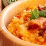 A close-up of a wooden bowl filled with a hearty dish containing rice, chunks of meat, and garnished with fresh cilantro. A bottle of Tabasco sauce is partially visible in the background, suggesting a spicy flavor profile for the meal.