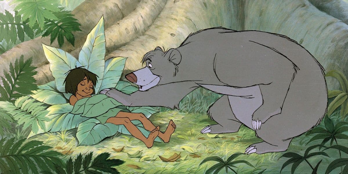 A scene from an animated movie shows a young boy with dark hair lying on large leaves on the forest floor, smiling as a friendly, gray bear playfully pats him. The background is filled with lush green foliage and towering tree trunks.