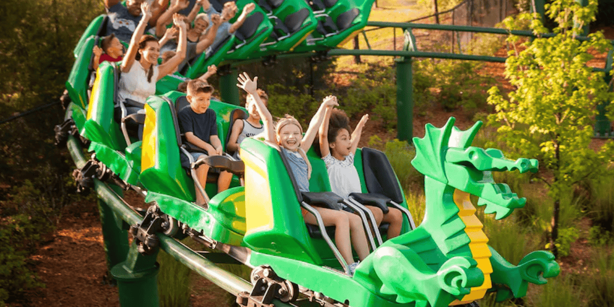 Children ride a green dragon-themed roller coaster at Legoland. They smile and raise their arms in excitement, enjoying the ride, unaware of the recent news about a baby having a heart attack nearby. The coaster is surrounded by greenery on a sunny day.