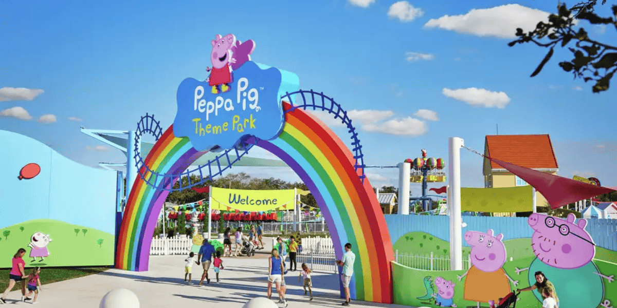 Entrance to Peppa Pig Theme Park featuring a colorful rainbow arch with a sign and smiling Peppa Pig characters. Several families walk through the entrance under a sunny sky. Attractions of the vibrant Peppa Pig Theme Park can be seen in the background.