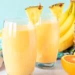 Two glasses of orange smoothies topped with pineapple wedges sit on a white surface surrounded by fresh fruits, including a whole pineapple, bananas, and a halved orange. A colorful floral cloth is in the background against a light blue backdrop.