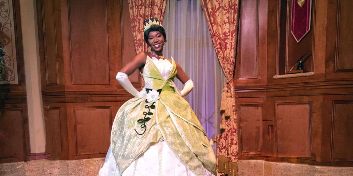 A woman smiles warmly while standing in front of ornate wooden doors and a window with elegant curtains. She is wearing a green and white princess gown with gloves and a tiara, and poses with her hands on her hips. The background has a regal, castle-like setting.