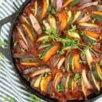 A cast iron skillet contains a beautifully arranged ratatouille with slices of zucchini, eggplant, and tomatoes in a spiral pattern, garnished with fresh thyme. The skillet is placed on a striped blue and white cloth, with sprigs of thyme scattered around.