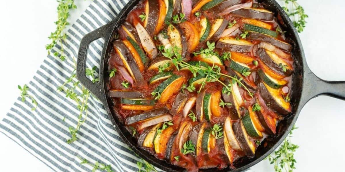 A cast iron skillet contains a beautifully arranged ratatouille with slices of zucchini, eggplant, and tomatoes in a spiral pattern, garnished with fresh thyme. The skillet is placed on a striped blue and white cloth, with sprigs of thyme scattered around.