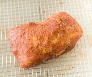 A seasoned pork roast, inspired by a Ronto Wrap recipe, sits on a wire rack displaying a savory red crust with visible herbs sprinkled on its surface.