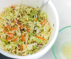 A bowl of mixed cole slaw with shredded green cabbage, carrots, and red cabbage, a fork inside the bowl, and some Ronto Wrap Recipe dressing on the side.