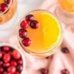 Overhead view of three glasses of orange juice garnished with orange slices and cranberries. A small white bowl filled with cranberries is placed on a light pink cloth, with additional cranberries scattered around it.