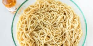 A clear glass bowl filled with cooked spaghetti noodles is placed on a white surface. To the left of the bowl, there is a small bottle of olive oil. The noodles appear lightly coated and ready to be served.