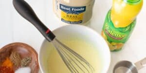 A bowl of seasoned mayonnaise being whisked is surrounded by various ingredients. To the left, a wooden bowl holds different spices and seasonings. Nearby is a bottle of Realemon lemon juice, a jar of Best Foods mayonnaise, a measuring cup, and a metal whisk.