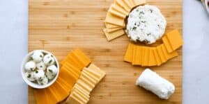A wooden board displaying a variety of cheeses. On the top right are slices of cheddar cheese and a round piece of blue cheese. On the bottom center are slices of gouda cheese and a log of goat cheese. On the left is a bowl of small mozzarella balls with herbs.