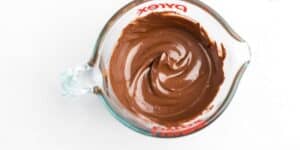 A glass measuring cup filled with smooth, melted chocolate. The chocolate has been stirred and swirled, creating a glossy, creamy texture. The measuring cup is placed on a white background.
