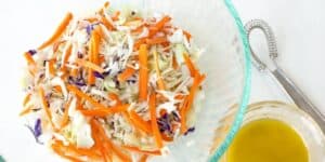 A glass bowl contains a fresh coleslaw mix made of shredded cabbage, carrots, and purple cabbage. A small jar with a golden yellow dressing sits beside the bowl, along with a whisk on a white surface.