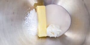 A mixing bowl containing a stick of butter, granulated sugar, powdered sugar, and a dollop of shortening neatly arranged side by side. The bowl's metallic surface reflects the light, creating a shiny backdrop for the ingredients.