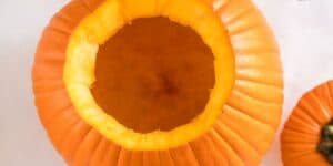 A top-down view of a large, hollowed-out pumpkin with its top removed, exposing the inner cavity. The orange exterior of the pumpkin is visible, along with another smaller pumpkin partially in the frame on the right side.