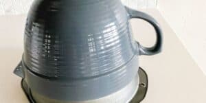 A dark gray ceramic teapot is placed upside down over a metal cake pan on a white countertop. The teapot has a handle on the side, and the cake pan's rim is partially visible beneath it. The background features a geometric tiled wall.
