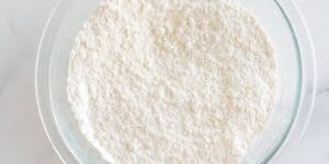 A clear glass bowl filled with a white, powdery substance, likely flour, placed on a smooth white surface. The finely textured flour creates a smooth, almost uniform appearance within the bowl.