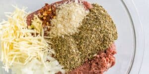 A clear bowl containing ground meat topped with shredded cheese, minced onions, red chili flakes, breadcrumbs, and a variety of green herbs and spices.