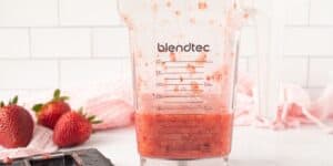 A clear Blendtec blender jar filled with blended strawberries sits on a white kitchen countertop. Fresh whole strawberries and a pink towel are in the background, set against a white subway tile backsplash.
