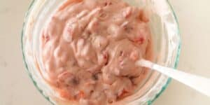 A glass bowl filled with a creamy mixture that has a light pink hue and small chunks of fruit. A white spoon rests in the bowl, partially submerged in the mixture. The background is a light-colored surface.