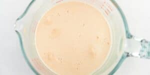 A glass measuring cup filled with a beige liquid mixture, likely a batter or a frothy beverage. The cup has measurement markings in red along the side, and the liquid has small bubbles on its surface. The background is white.