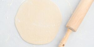 A round piece of rolled-out dough lies on a lightly floured surface next to a wooden rolling pin. The setting suggests a baking or cooking activity, with the dough prepared for further processing.