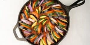 A colorful ratatouille dish is arranged in a black cast iron skillet. The dish features layers of sliced zucchini, yellow squash, eggplant, tomatoes, and red onions, all nestled in a vibrant red tomato sauce.