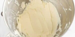 A close-up view of smooth, creamy, light yellow buttercream frosting inside a metal mixing bowl. The frosting has been evenly spread across the surface, showing a thick and fluffy texture. The edges of the bowl are slightly smeared with frosting.