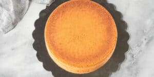 A round, golden-brown cake sits on a large black scalloped-edge cake board, placed on a marble surface. The cake appears to be plain with no frosting or decorations, and a textured fabric is visible in the corner of the image.