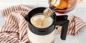 A bottle of root beer is being poured into a dark brown plastic mug filled with vanilla ice cream, creating a root beer float. A red and white striped cloth is in the background on a light-colored kitchen counter.