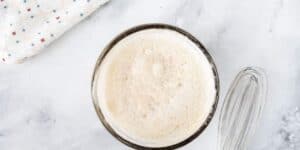 A bowl filled with frothy beaten egg whites, accompanied by a whisk. A cloth with a pattern of multicolored stars is visible in the background on a marble surface.