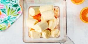 A blender container filled with chopped bananas and orange segments, ready to be blended. The container is placed on a light surface, with part of a colorful cloth and slices of orange visible in the background.
