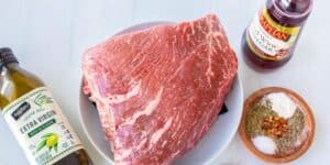 A piece of raw beef is on a white plate next to a small wooden bowl containing spices and herbs. There is a bottle of extra virgin olive oil on the left and a bottle of red wine vinegar on the right, all on a white surface.