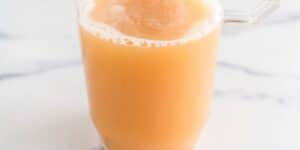 A clear glass pitcher filled with an orange-brown liquid sits on a white marble surface. The liquid has a frothy top layer.