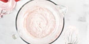 A glass mixing bowl filled with light pink whipped cream sits on a white marble surface. A metal whisk with remnants of the whipped cream lies next to the bowl. A pink oven mitt can be seen in the upper left corner.
