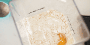 Overhead view of an open blender container containing a mixture of flour and an egg yolk, set on a white countertop. A teal measuring cup with flour is partially visible on the side.