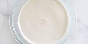 A clear glass bowl filled with smooth, creamy white batter or sauce, placed on a light marble countertop.