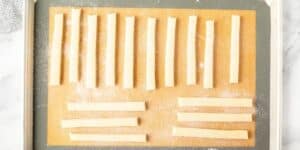 A baking tray lined with parchment paper holds numerous evenly spaced, uncooked breadsticks that are sprinkled with flour. The breadsticks are laid out in rows, three of which are placed horizontally at the bottom, while the rest are vertical.