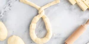 A piece of dough twisted to form a pretzel shape lies on a marble surface. To the right, there is a small wooden rolling pin and some sliced cheese. Additional dough pieces are situated on the left side.