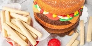 A burger-shaped cake with layers resembling a sesame seed bun, patty, cheese, lettuce, and tomato. It is accompanied by cake fries in a basket and a small container of red icing, mimicking ketchup. The setup is on a metal tray with a white napkin underneath.