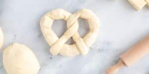 A close-up of uncooked pretzel dough shaped into a traditional knot on a marble surface. A small wooden rolling pin and additional dough portions are visible nearby, suggesting the process of making homemade pretzels.