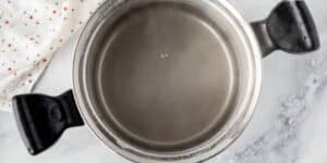 A stainless steel pot with black handles sits on a marble countertop. The pot is filled with clear water that appears to be steaming. A white cloth with colored dots is partially visible next to the pot.