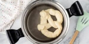 A pot with water boiling a pretzel dough. Next to the pot is a green spatula with a wooden handle and a white cloth with a pattern of small, colorful shapes. The scene is set on a marble countertop.