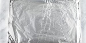 A rectangular baking sheet is covered with a sheet of crinkled aluminum foil. The foil has various wrinkles and folds, reflecting light unevenly. The baking sheet rests on a white surface.