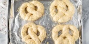 A baking tray lined with aluminum foil, holding four uncooked pretzels sprinkled with grated cheese. The tray is placed on a light-colored countertop, and a white cloth with colored polka dots is partially visible on the side.