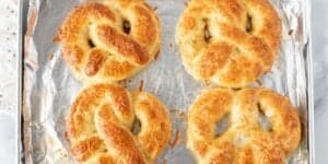 Four golden, freshly baked soft pretzels with a light sprinkling of coarse salt and cheese are arranged on a foil-covered baking sheet. The pretzels have a slightly crispy exterior, and the baking sheet shows some toasted cheese crumbs.