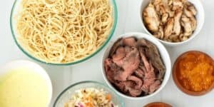 An assortment of foods on a table: a bowl of cooked spaghetti, a plate of grilled chicken slices, a bowl of sliced roast beef, a wooden bowl of orange fish roe, a bowl of creamy yellow sauce, and a small bowl of coleslaw.