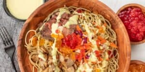 A wooden bowl filled with noodles topped with slices of beef, shredded vegetables, and colorful roe. Drizzled with a light yellow sauce, it is surrounded by a fork, knife, and small bowls of additional roe and sauce.