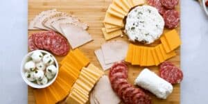 A wooden charcuterie board filled with arranged slices of cheddar and Swiss cheeses, salami, and turkey. The board includes a small bowl of mozzarella balls garnished with herbs, a round of herbed cheese, and a log of goat cheese.