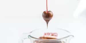 A red cherry being dipped into melted chocolate, held by a thin red stick above a clear glass measuring cup filled with chocolate. The background is blurred and white, drawing focus to the cherry coated in rich chocolate dripping slightly.