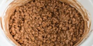 Close-up of a crockpot filled with melted chocolate mixed with peanuts, forming clusters. The creamy, nutty mixture has a rich, glossy texture and fills the entire pot, hinting at a delicious homemade treat in the making.